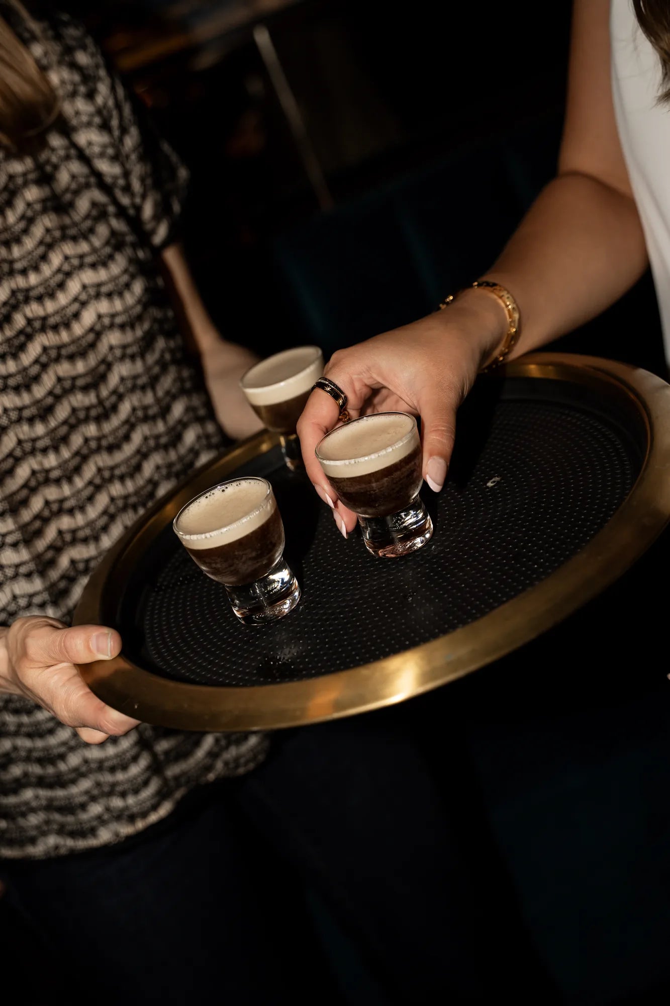 A woman holding a gold and black tray of espresso martini shots at a cocktail party with Royce + Reign event favor cookies.